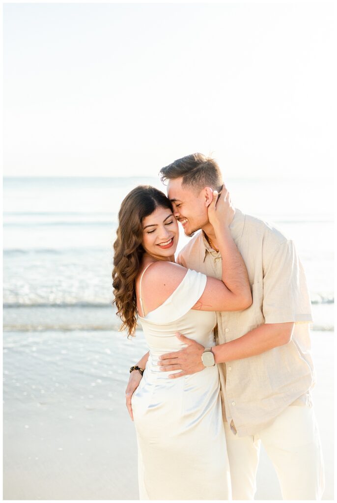 A couple named Julissa and Dallas, newly engaged, embrace each other lovingly. Dallas' nose is at Julissa's temple as he smiles right before kissing her temple and she is giggling down her shoulder as he does this. It's a a close shot of them with the calm Puerto Rico ocean behind them.