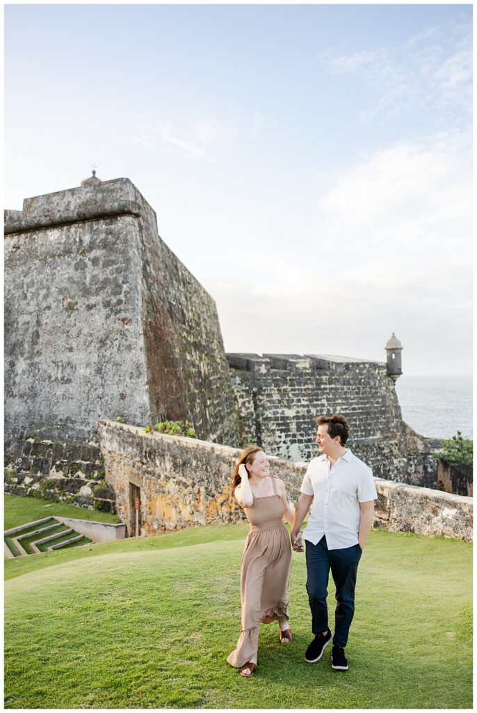 A newly engaged couple walking together during their engagement session in Puerto Rico. Their backdrop is the El Morro Fort in Old San Juan.