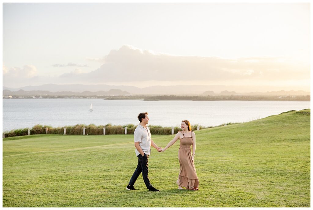 Couple walking together during an engagement session in Puerto Rico at sunset. Their backdrop shows the green grass at El Morro with the ocean in the background.