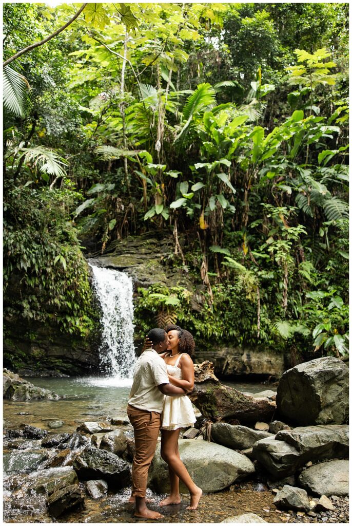 A newly engaged couple embracing and kissing in front of a romantic waterfall at the El Yunque Rainforest in Puerto Rico.