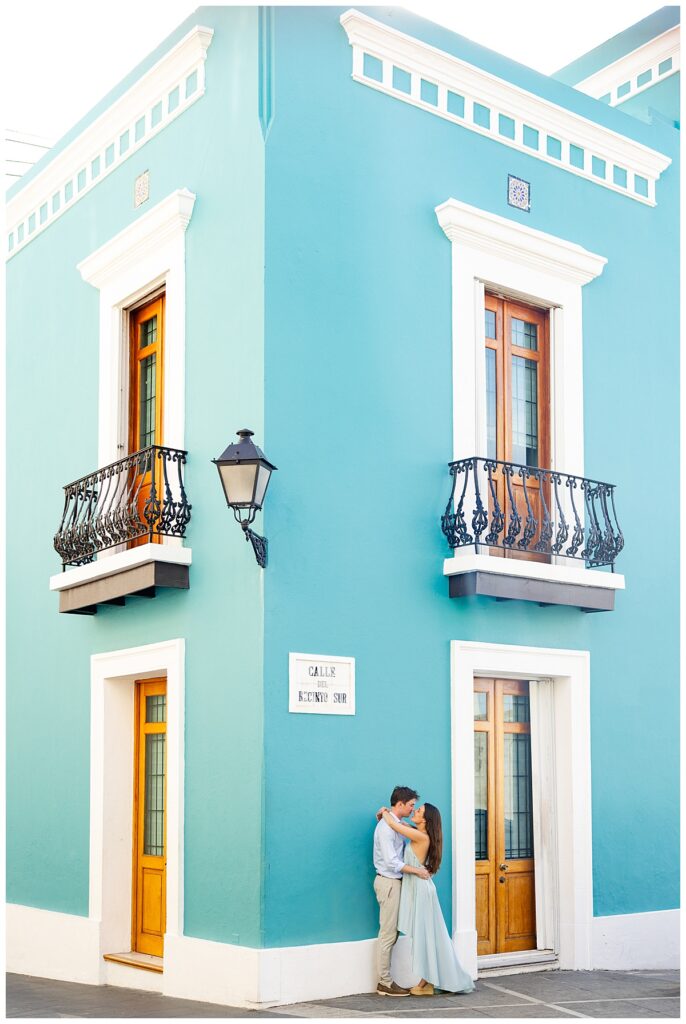 A couple hugging each other with a bright blue Old San Juan home as their background. Session in Old San Juan