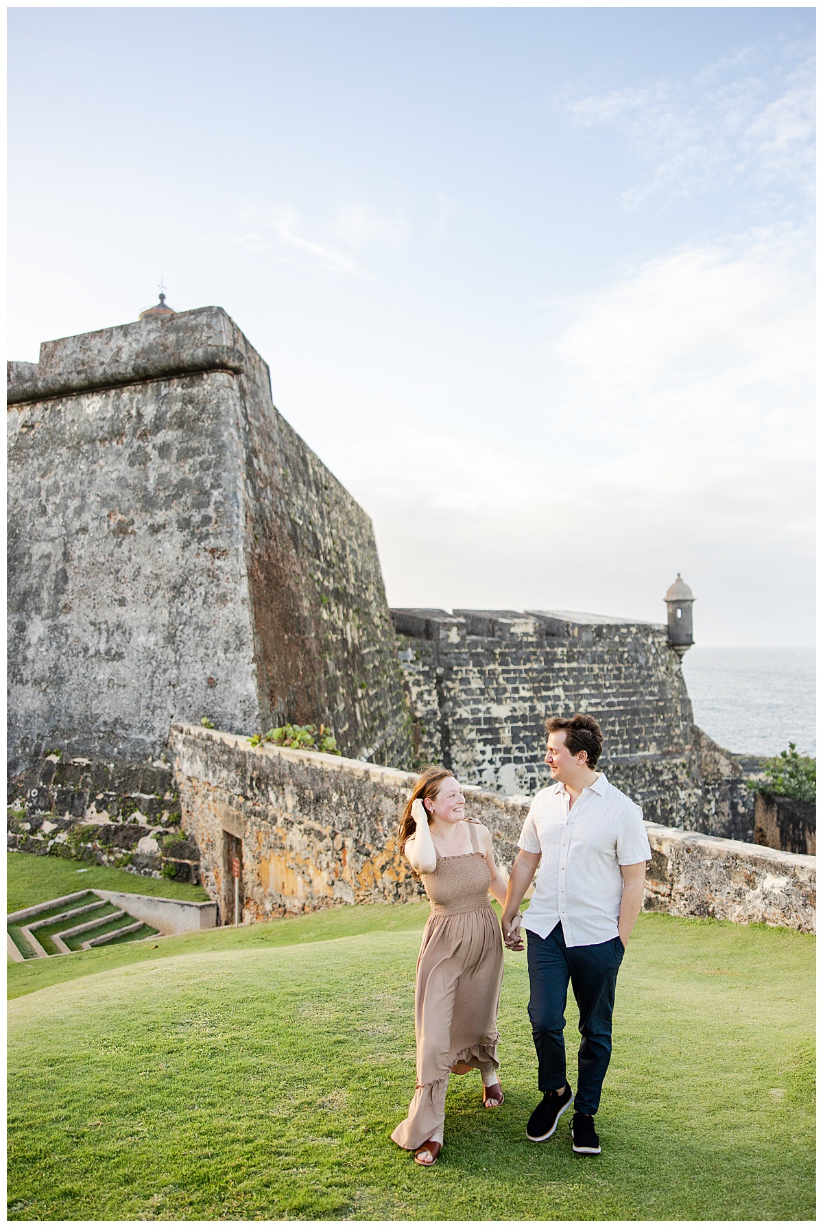 A newly engaged couple walking together during their engagement session in Puerto Rico. Their backdrop is the El Morro Fort in Old San Juan.